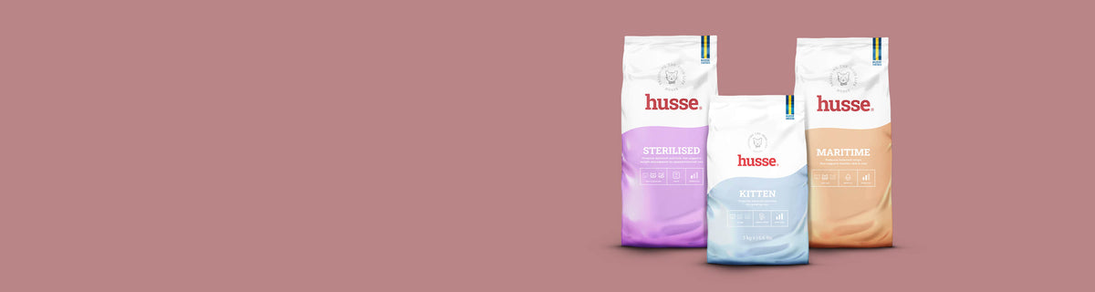 Three bags from the Husse Essential cat food range on a dusty pink background